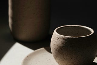 Three stoneware articles — plate, cup, and jug — stand on a surface. The stoneware articles and surface are partly lit by sunlight.