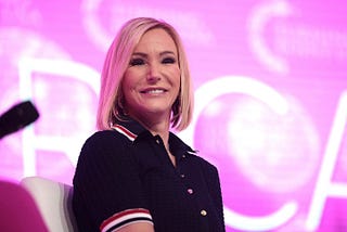 A middle-=aged woman with shoulder-length straight blonde hair, wearing a button-down shirt dress with red, white, and blue ribbons at the collar and sleeves, smiles down from a stage.