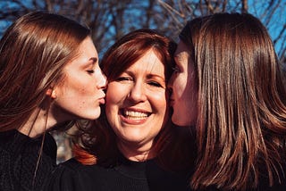 Two brown haired girls kissing their mom on the cheeks