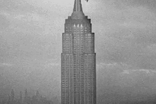 Long shot of the top half of the Empire State Building against a cloudy sky. King Kong falls from the very top, bouncing on the way down.