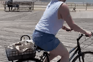 A cyclist rides her bike down a wide boardwalk on a sunny day, the ocean visible in the background.