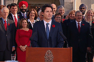 Justin Trudeau shrugging at a press conference. He is standing in front of the members of his cabinet from 2015.