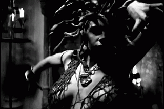 A gif of a woman, meant to represent Medusa. They have snakes for hair, and are dancing sensually in front of a large fireplace in a tattered robe.