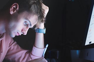 A man at his desk in the dark. He is in front of a computer while looking down at his iPad and holding an Apple Pencil.