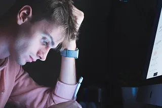 A man at his desk in the dark. He is in front of a computer while looking down at his iPad and holding an Apple Pencil.