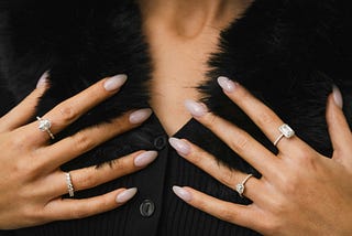 A woman, the image cropped as she displays multiple rings on her hands against a fur coat.