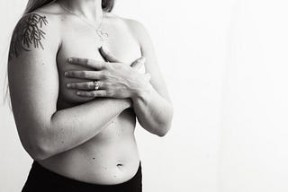 In black and white, a lady naked from waist up, holding her breasts to hide or protect them.