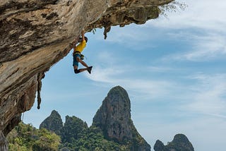 A rock climber holds on (leggs hanging) to a sheer rock face above blue water with verdant islands in the background.