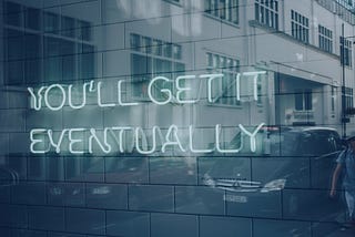 A storefront window with a blue neon sign that reads, “You’ll Get it Eventually, reflecting a lone figure on a narrow street