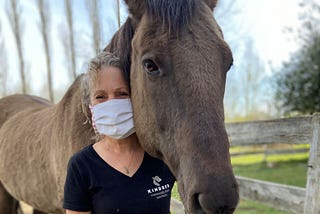 Keryn Denroche, a woman with short gray hair and a gray mask, poses with a soft looking brown horse on her farm sanctuary