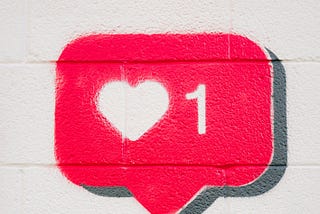 A hand holding a smartphone under a painting of a social media heart “love” symbol.