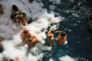 Suntanned women and men having a pool party. They are drinking cocktails and the pool is covered with foam.