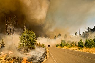 A vehicle on a road driving away from a very smoky wildfire in the background.