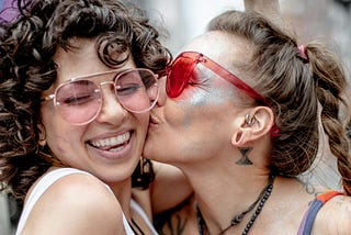 One woman laughing while a second woman kisses her on the cheek. They are wearing rainbow attire and are both smiling.