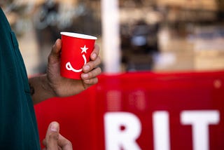 A hand holding a red to-go coffee cup with Ritual’s logo—a curved line signifying a mug below a star.