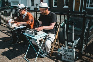Two men writing on typewriters in the park. There is a fence and a road behind them. One man is wearing white pants, a black shirt, and a hat. The second man is wearing black pants, an orange shirt, a scarf, and a hat.