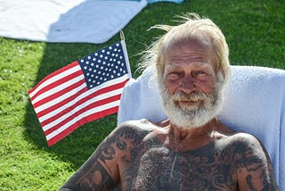 Tattooed man sitting shirtless with American flag behind him.