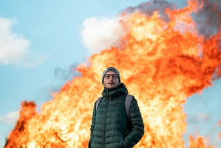 Man in green jacket dark pants, hands in pocket, wearing a grey beanie, standing in front of explosion fire.