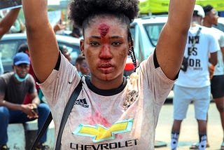 Attractive young woman holds aloft a sign saying, “EndPoliceBrutality” during a protest. She is costumed like a victim.