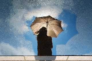 Woman looking at her reflection on a sidewalk, but instead of her reflection, she sees a painting instead. In the painting, she’s holding an open umbrella under a blue sky full of white clouds and what seems to be stars.