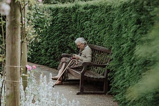 A woman sits on a wooden bench next to a hedge in a garden.