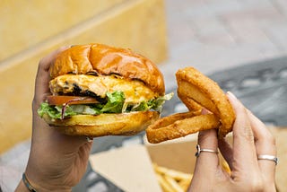 A woman sits at a table, a burger in one hand, three onion rings in the other. A basket of french fries sits on the table.