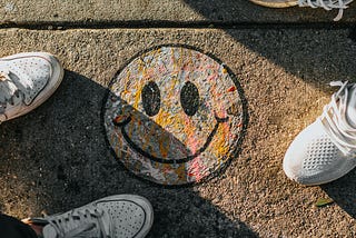 A smiley face that has been chalked onto the sidewalk, with the sneakers of people around it. Photo taken from above.
