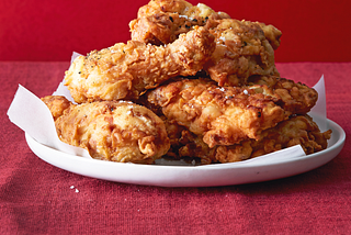 A white dish with a heap of fried chicken parts, on a red tablecloth with a red background.