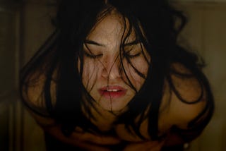 Woman with dark hair and closed eyes, leans the back of her head on the glass shower door.