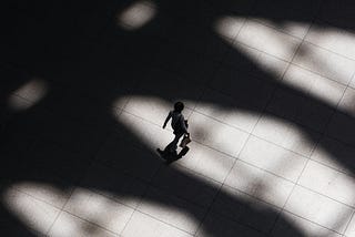 In a black and white photo, a person is seen from a bird’s eye view walking through a scene in which large shadows are cast from an unknown source.