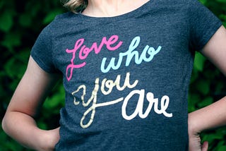 Young person standing proudly with hands on hips, looking upwards, wearing a T-shirt saying ‘Love who you are’