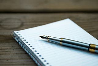 An empty notebook sits on a wooden desk, with a pen sitting atop it.