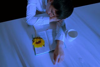 A man sleeping on a desk with an open book, a sunflower, and a white cup with some spilled water in front of him