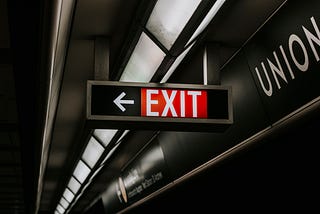 Exit sign with white lettering on a red background, suspended from the ceiling of a public room