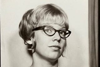 Headshot of author at 22, wearing horn rim glasses, sleeveless dress, and an updo hairstyle with bangs