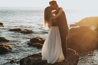 A tall man romantically clasps in his arms a long-haired woman in a flowing white dress. They’re kissing while standing on a rock on the beach, surrounded by water and mistiness to the horizon.