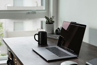 A home office setup. A desk sits next to a window. It has a laptop with a mouse, a coffee cup, a book upon which sits a plant and a camera, and other knickknacks. A pair of headphones hangs from one of the desk legs.