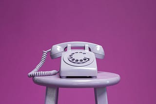 Rotary phone on a barstool with a pink background