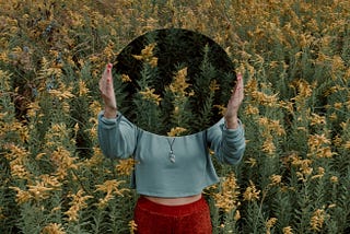 A woman standing in a field of wildflowers, holding up a mirror to cover her face.