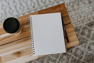 A blank notebook, a pen, and a cup of coffee sit on a wooden desk.