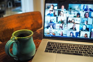 A green coffee mug placed next to an open laptop, with a mosaic of faces in the middle of a video conference call.
