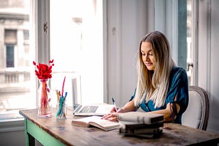 A female sits at their desk, writing in a journal with their laptop and pens nearby. #writing #journaling #writer