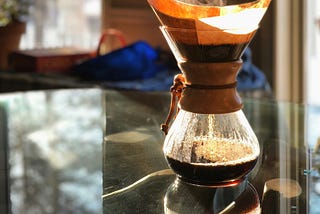 A Chemex coffee maker brewing coffee, with steam rising from the top. The warm sunlight illuminates the scene, casting reflections on the glass table. A blurred background with greenery and a framed picture add a cozy atmosphere.