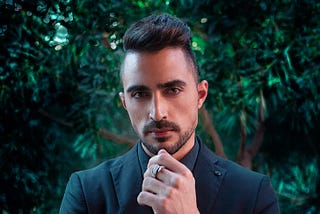 Sleek dressed male with side cuts hairstyle and trimmed beard, wearing blazer, watch, looking into camera determined.