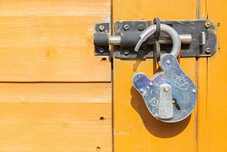 A heavy-duty silver padlock hanging completely open and unlatched from a dark metal slide bolt lock on a bright, sunlit yellow wooden door. The open lock sits on the right side of the horizontal frame, symbolizing freedom, broken silence, and midlife liberation