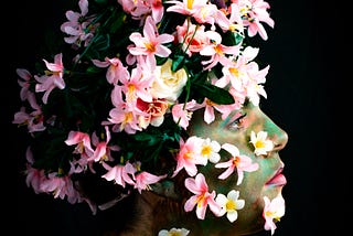 A woman with a pensive, somewhat mournful expression poses with her face, hair and upper body covered in flowers and her skin painted earthy green so that she is a living garden.