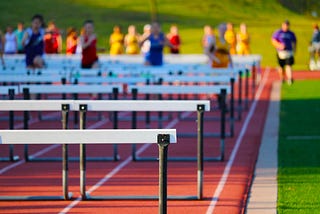 Adolescent children in colorful pennies run on a track with hurdles. The hurdles are in the foreground on a red/brown track and the kids are positioned toward the back of the photo, out of focus. They were pennies that are blue, yellow, red, and purple, and they appear to be running toward the camera.