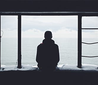 the silhouette of a man (alone) looking out a window to the sea