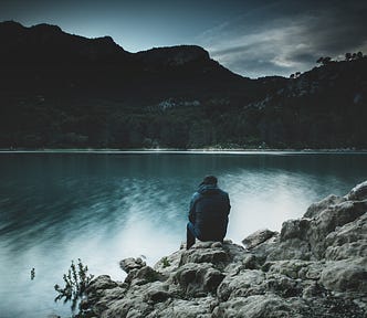 Image of a man sitting on the rocks on a lakeside. Image is dark and gloomy signifying loss, endurance and time to heal.