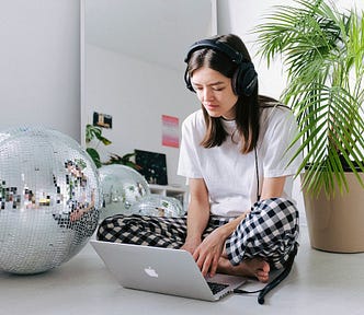 Woman in white shirt and pajamas using MacBook next to a disco ball and plant